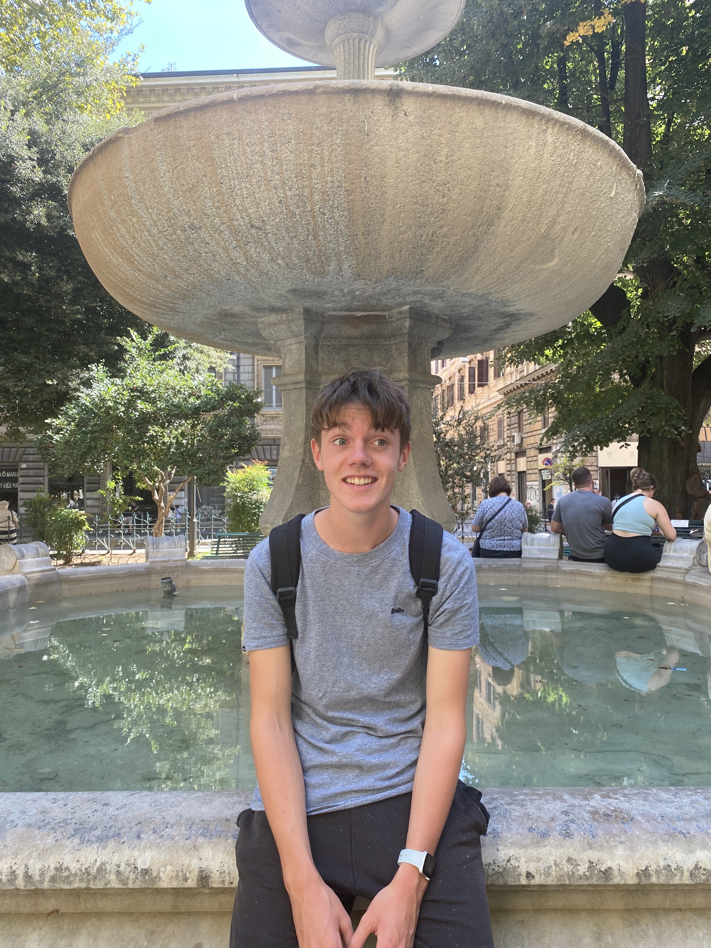 Olly smiling, sitting at the border of a stone fountain. He is looking to the right of the camera, and there are a few people sitting on the other side of the fountain border.