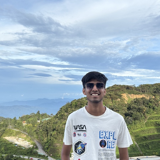 Pranav smiling broadly at the camera, wearing sunglasses and a white NASA t-shirt. The background shows a partly clouded blue sky and green hills.